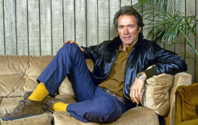 Clint Eastwood sits casually on a beige couch, wearing a black leather jacket, brown shirt, blue jeans, yellow socks, and brown shoes. The wooden wall and a green plant are in the background.