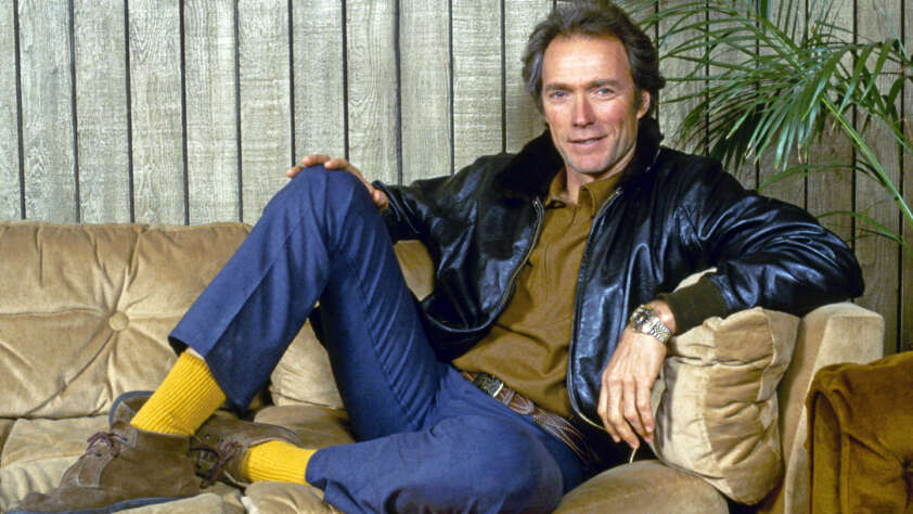 Clint Eastwood sits casually on a beige couch, wearing a black leather jacket, brown shirt, blue jeans, yellow socks, and brown shoes. The wooden wall and a green plant are in the background.