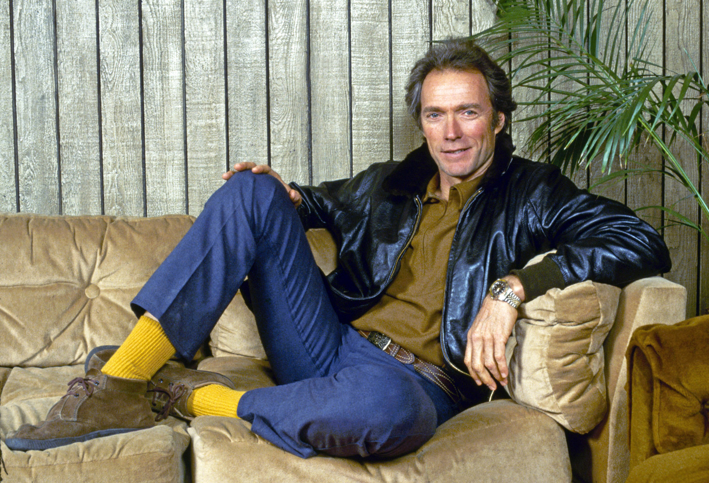 Clint Eastwood sits casually on a beige couch, wearing a black leather jacket, brown shirt, blue jeans, yellow socks, and brown shoes. The wooden wall and a green plant are in the background.